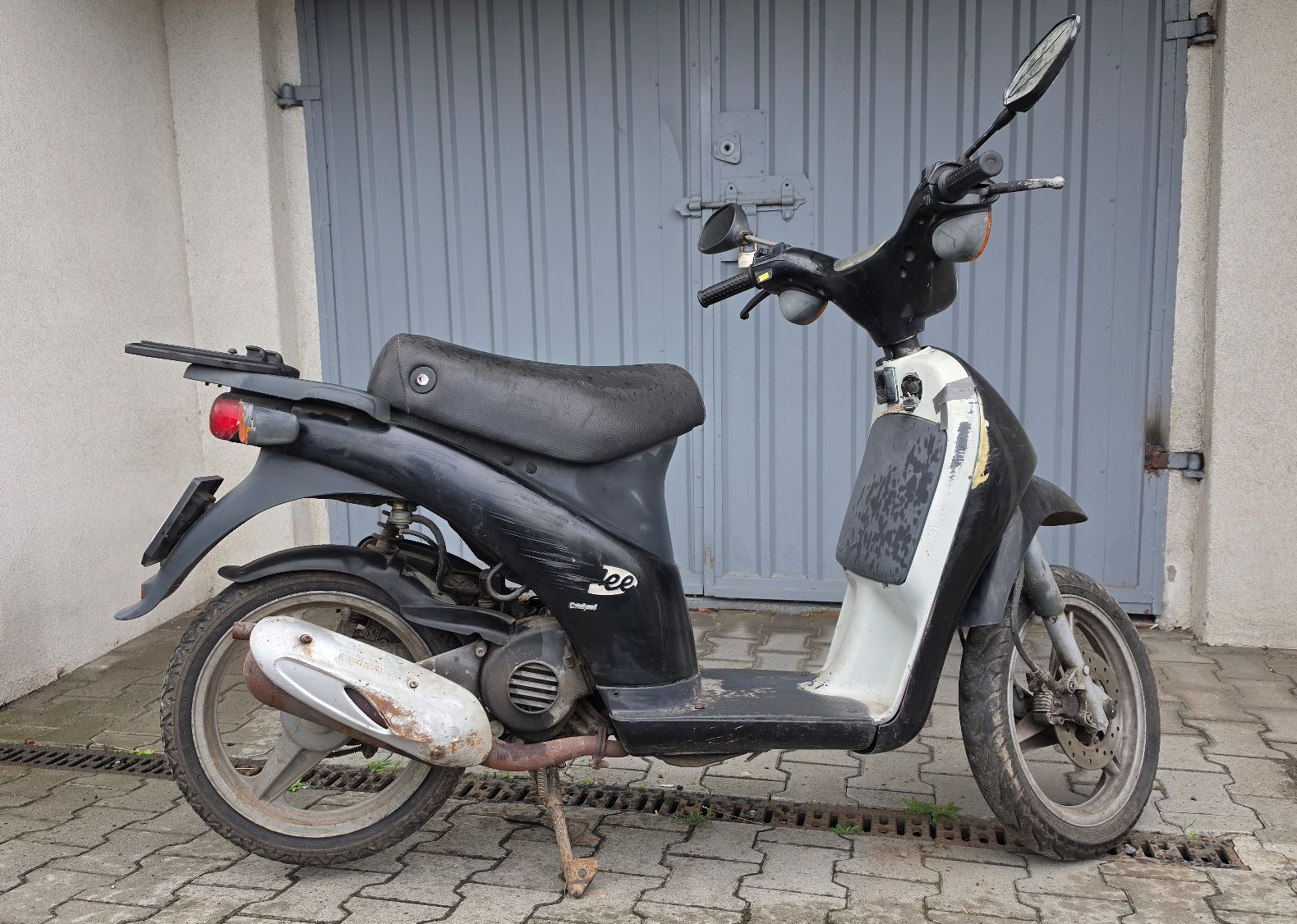 Motorower koloru czarnego. Widok z prawego boku. Ślady korozji na osłonie rury wydechowej. Siedzisko dla jednej osoby.