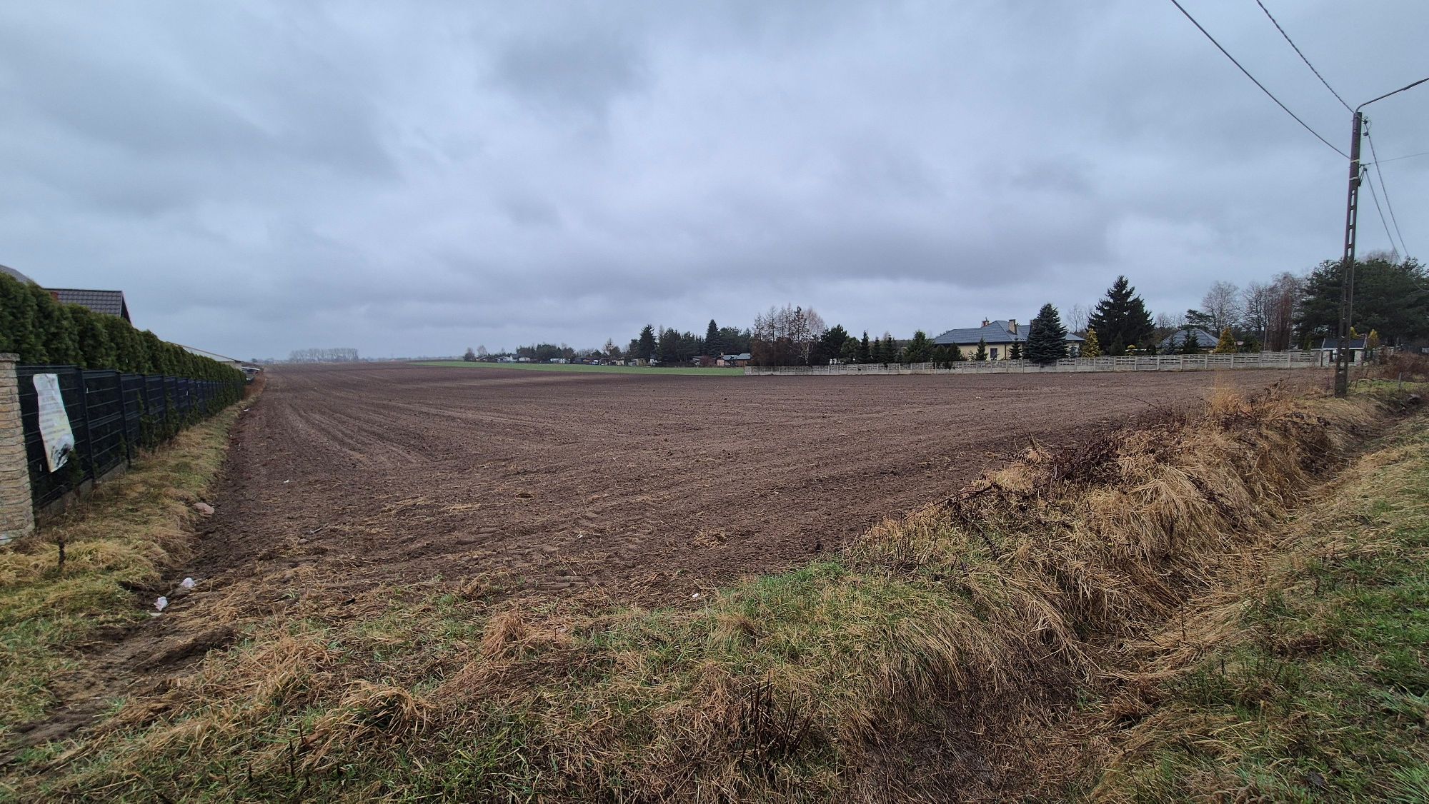 Puste, przeorane pole uprawne graniczące z ogrodzeniem i przydrożnym rowem pod zachmurzonym niebem.