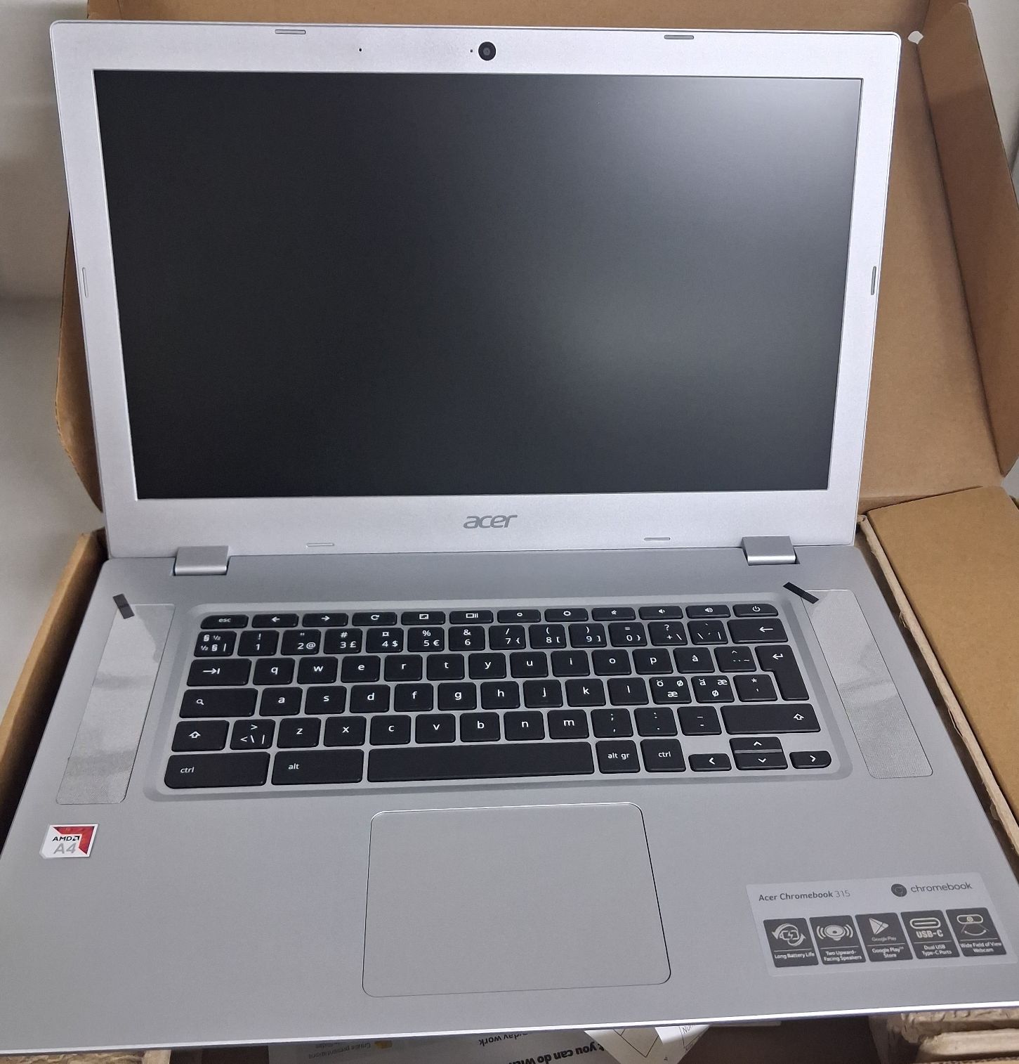 Laptop koloru srebrnego z widoczną klawiaturą i ekranem w opakowaniu kartonowym koloru brązowego.