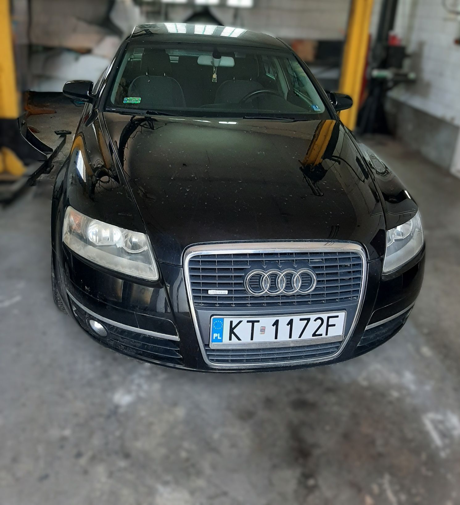 Zdjęcie przedstawia czarne Audi A6. Z przodu jest atrapa chłodnicy z chromowaną obwódką z logo. Podłużne klosze reflektorów