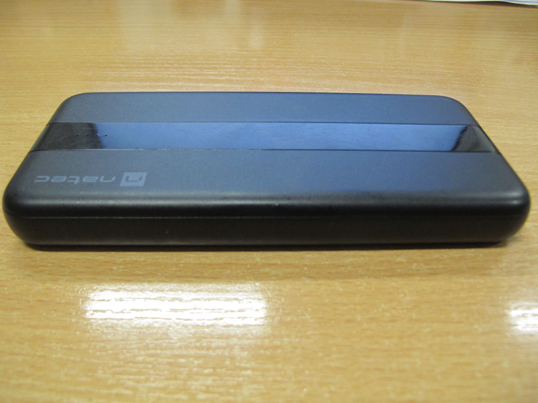 Czarny powerbank Natec leżący poziomo na drewnianym stole. Widoczne logo marki.