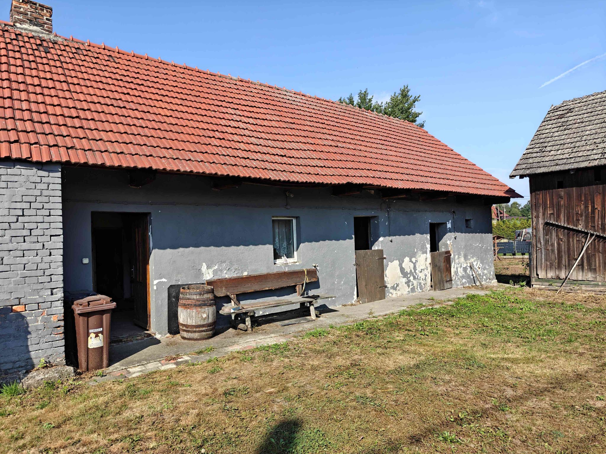 Widok na zabudowania gospodarcze. Po lewej stronie murowany budynek z jasnoszarą elewacją a po prawej drewniana stodoła.