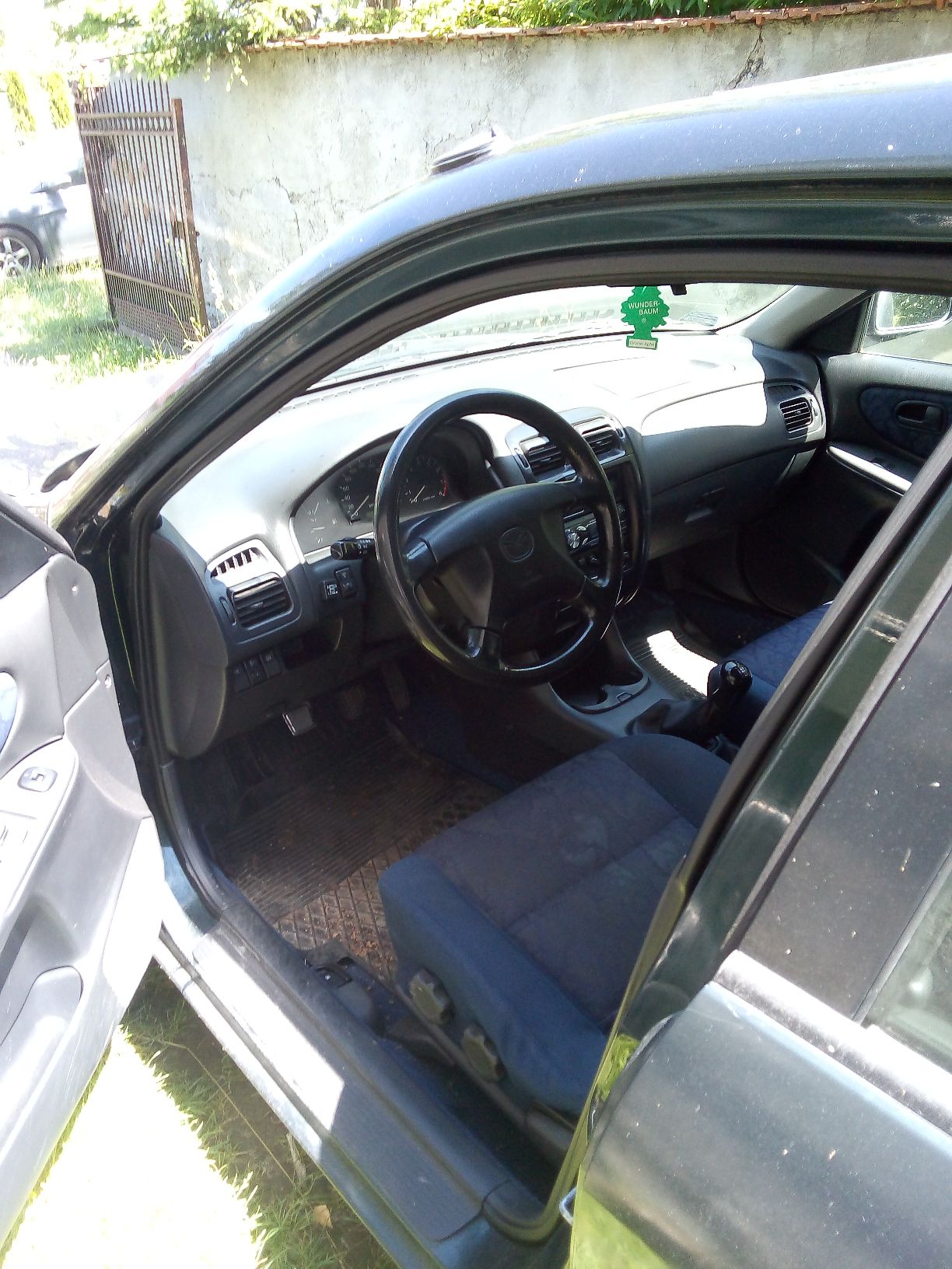 Wnętrze ciemnozielonego samochodu osobowego marki Mazda 626 widziane przez otwarte drzwi kierowcy z deską rozdzielczą i fotelami