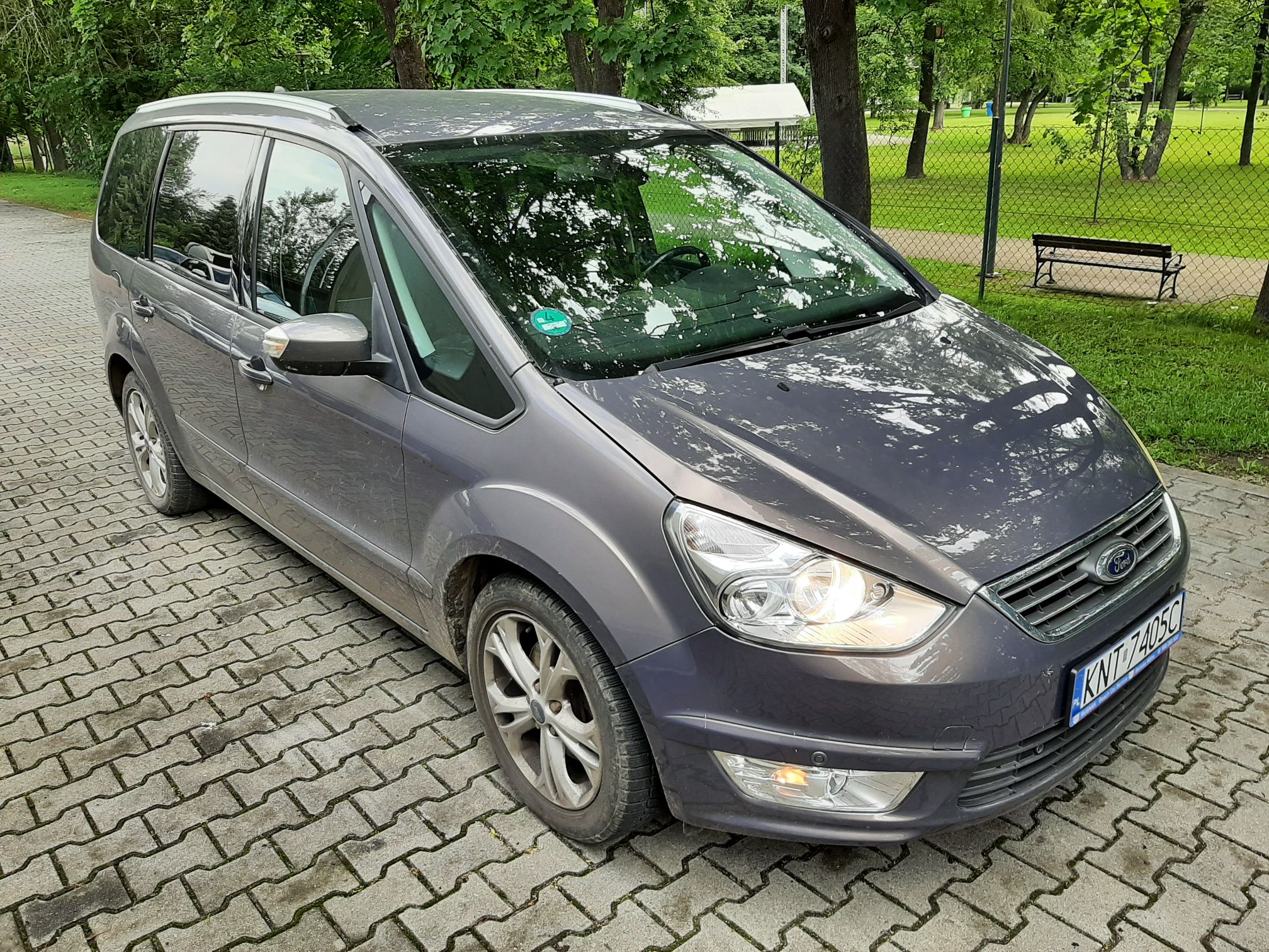Samochód osobowy marki Ford, szary, posiada reflektory włączone oraz widoczne tablice rejestracyjne z numerem.
