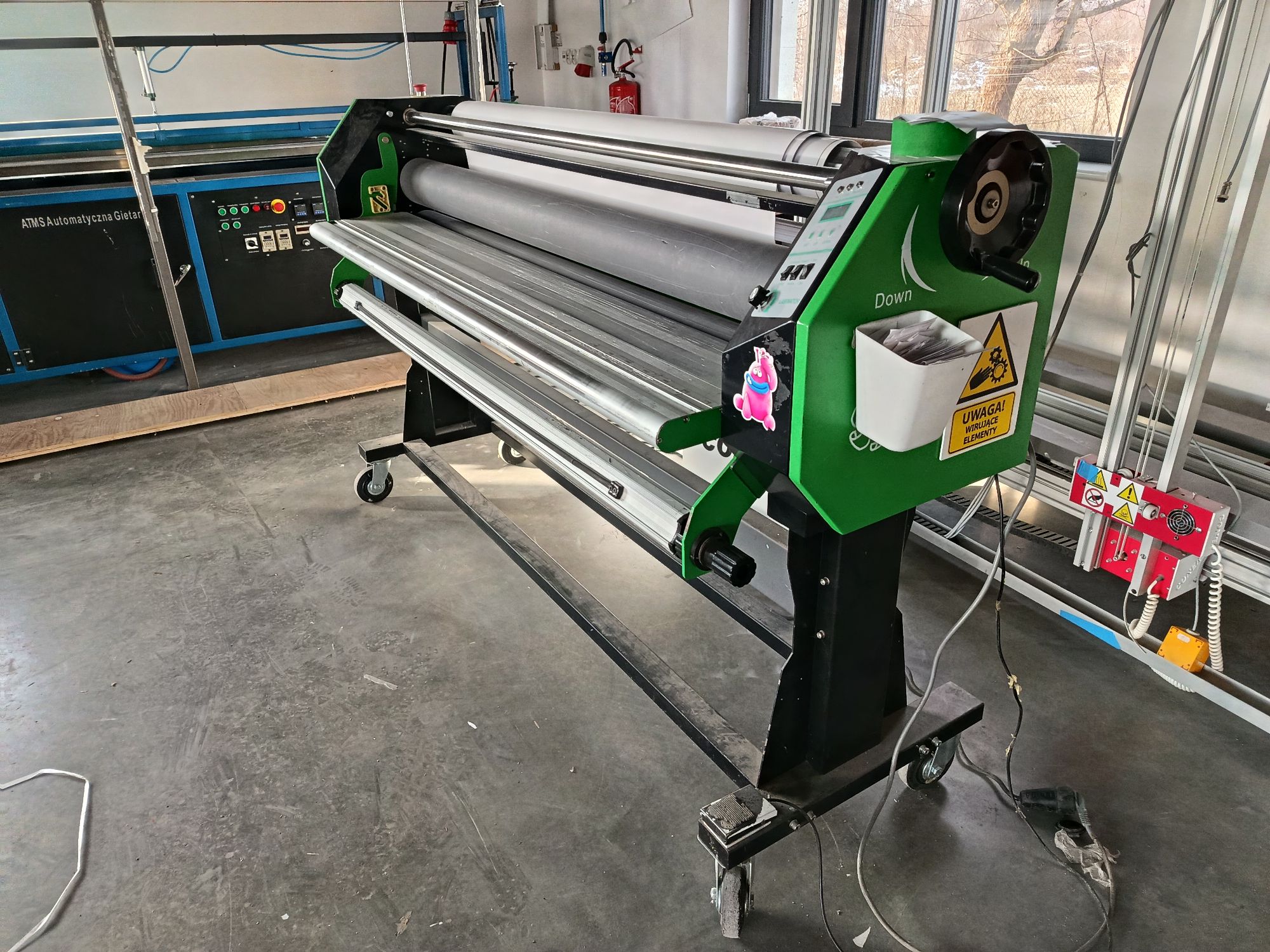 Wielkoformatowy laminator rolowy w kolorze zielono-czarnym, stojący na kołkach w warsztacie lub drukarni.
