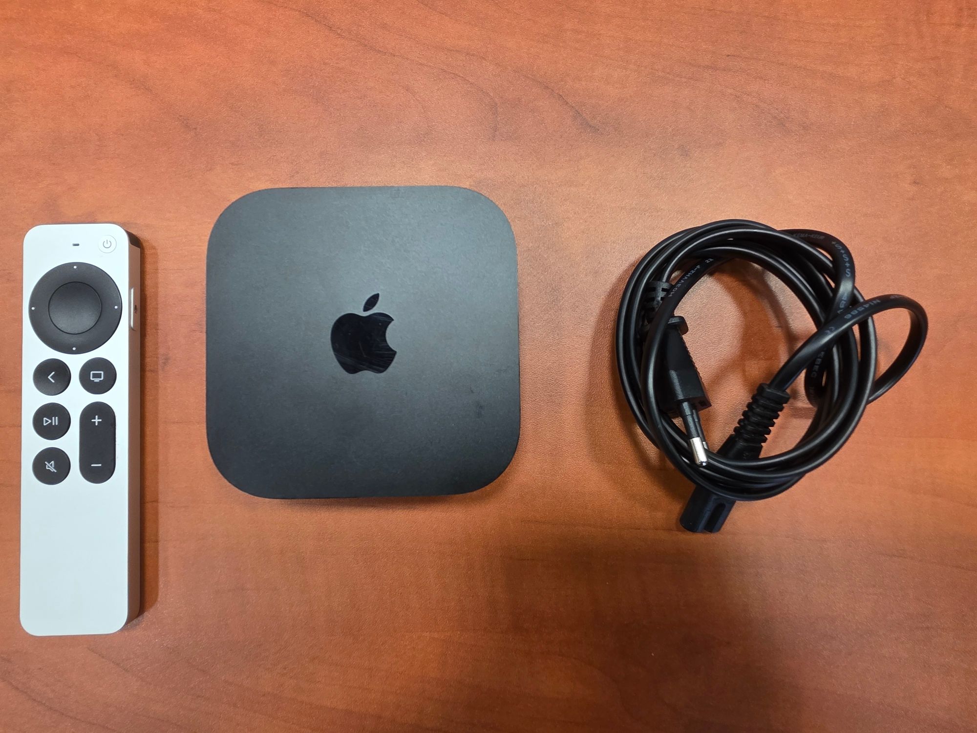 Urządzenie Apple TV koloru czarnego wraz ze srebrnym pilotem i czarnym kablem zasilającym