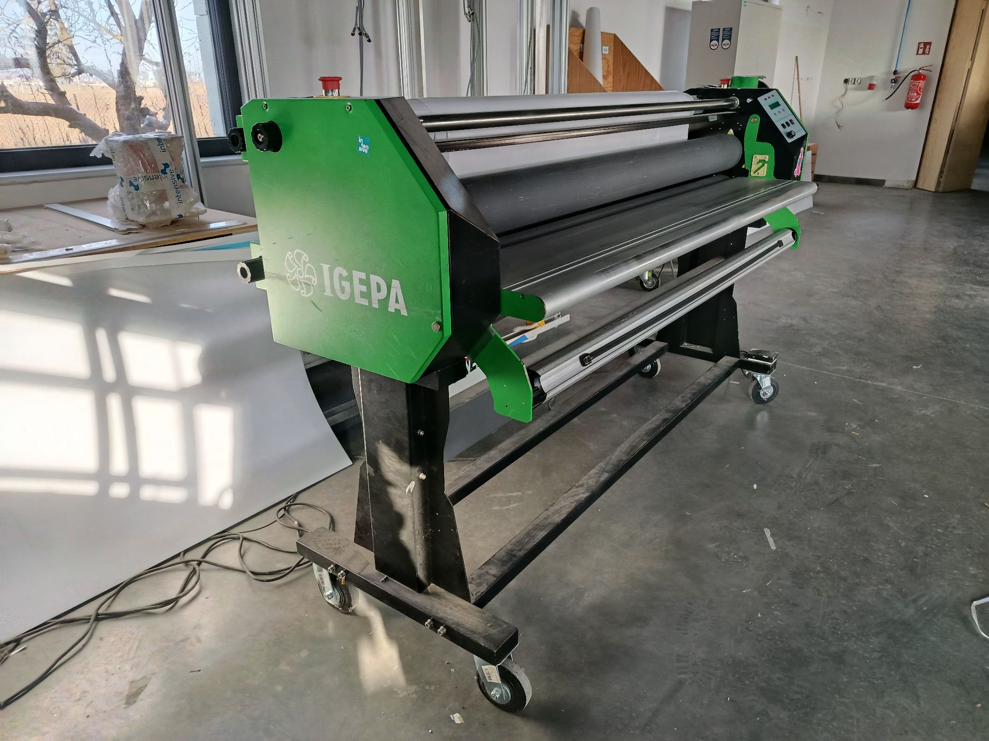 Wielkoformatowy laminator rolowy w kolorze zielono-czarnym, stojący na kołkach w warsztacie lub drukarni.