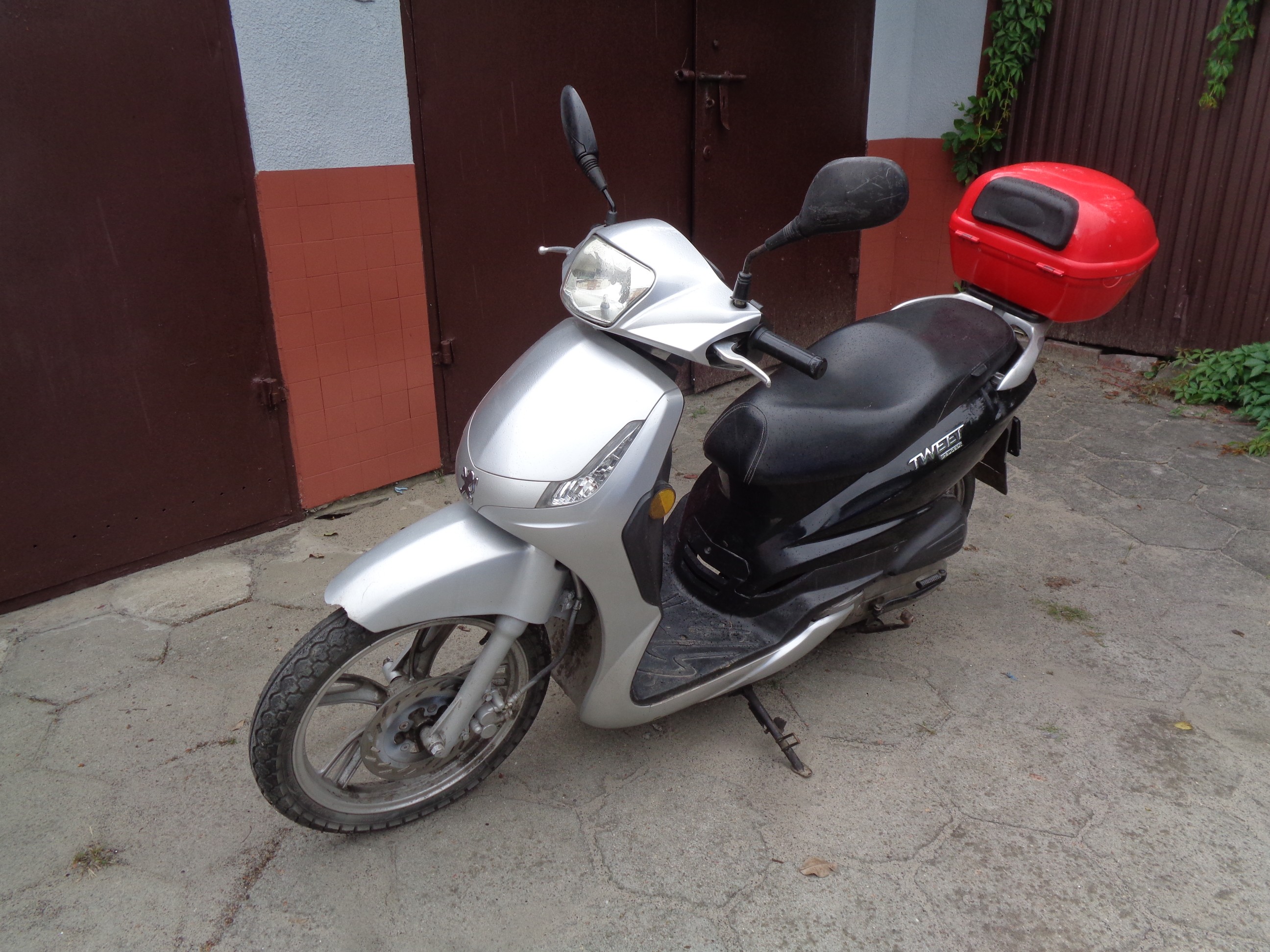 Srebrny motorower Peugeot z czarnym siedzeniem, widoczny z profilu, z tyłu zamontowany czerwony kufer. Lekkie zarysowania.