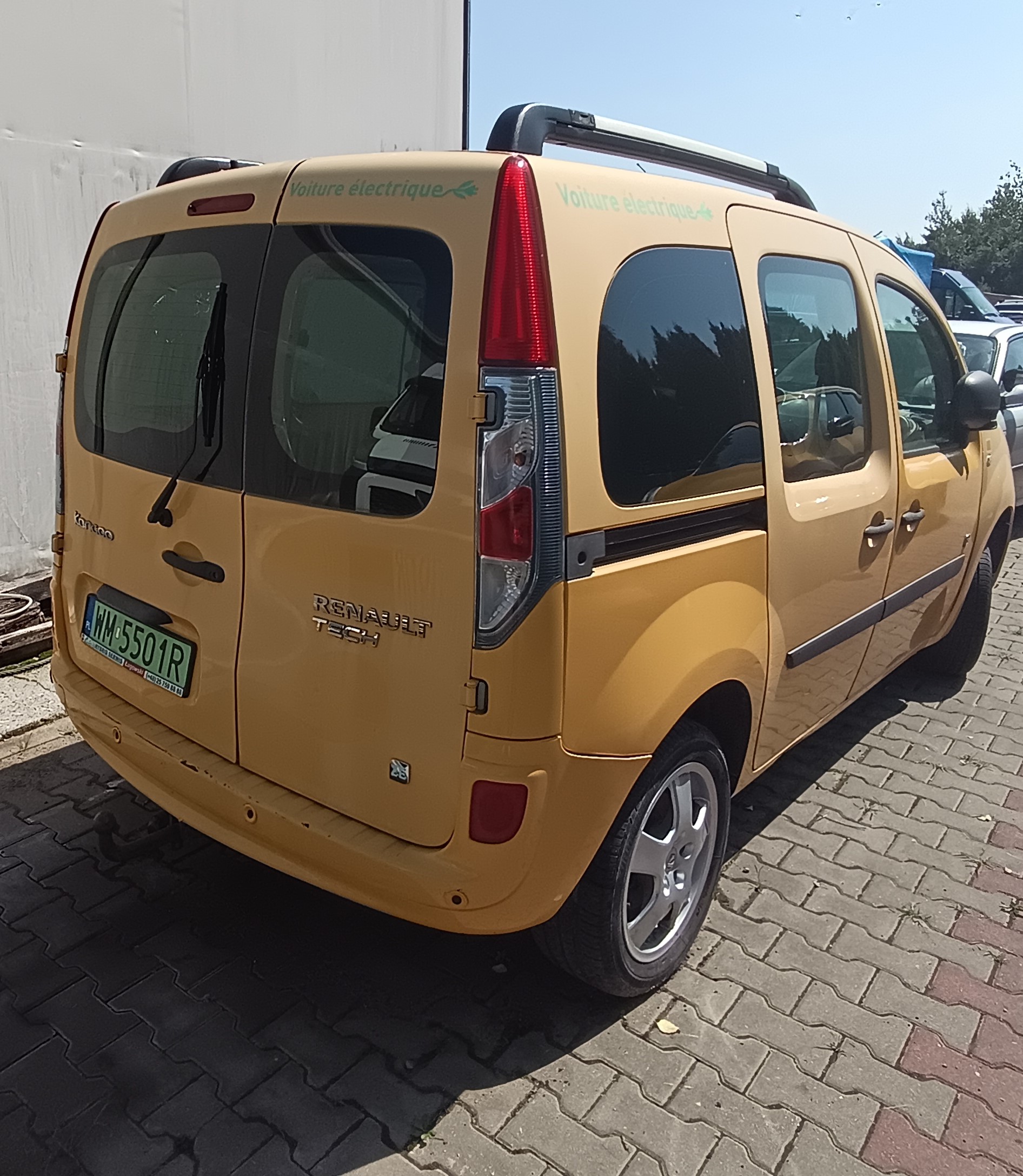 Żółty samochód dostawczy Renault Kangoo widziany z tyłu pod kątem, z dwuskrzydłowymi tylnymi drzwiami i pionowymi lampami.