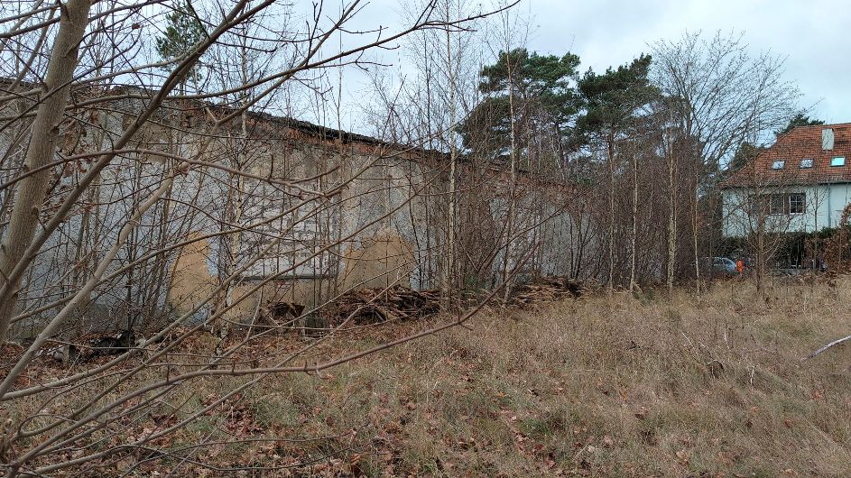 Trawa i gołe drzewa otaczają stary, zwietrzały betonowy budynek obok domu mieszkalnego