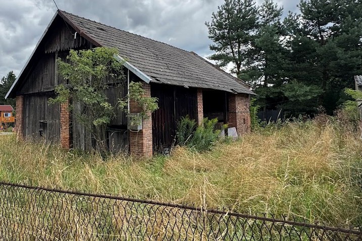 widok na stary drewniany budynek pod kątem z prawej strony. Z przodu na dole widoczny fragment ogrodzenia z metalowej siatki