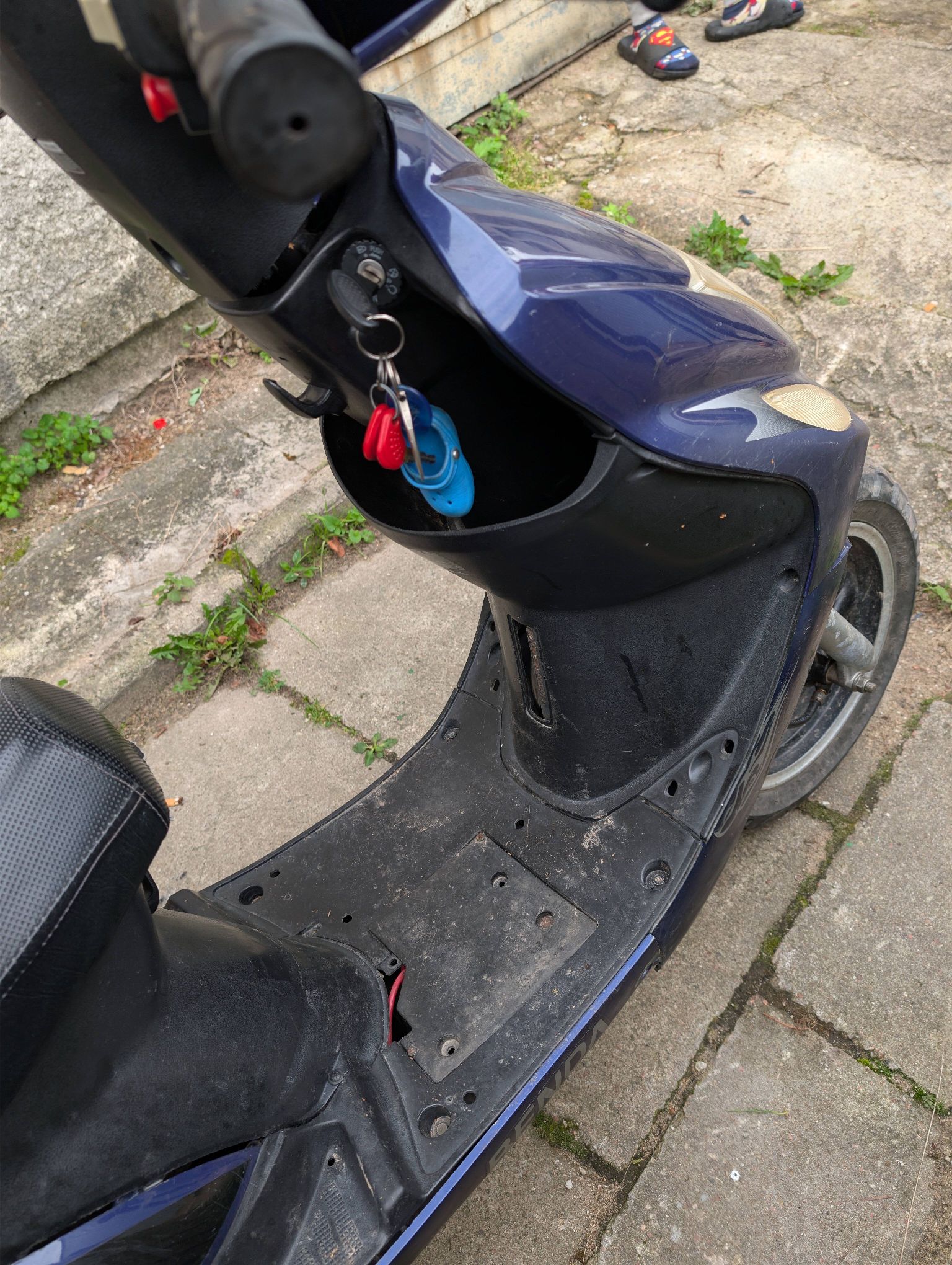 Niebieski motorower, sfotografowany z góry. Widoczny czarny, plastikowy podest na nogi, który nosi ślady użytkowania.