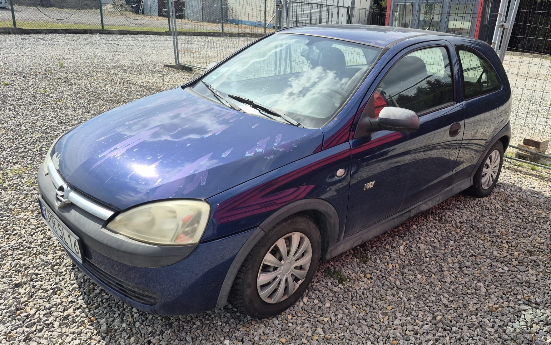 Lewa strona granatowego pojazdu OPEL CORSA C. Ubytki lakieru na masce, plastikowe nakładki na progi oraz srebrne kołpaki.