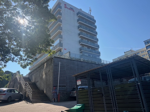 Apartamentowiec nr 34, betonowe schody prowadzące na podwyższenie, na dole parking samochodowy i zadaszona wiata śmietnikowa.