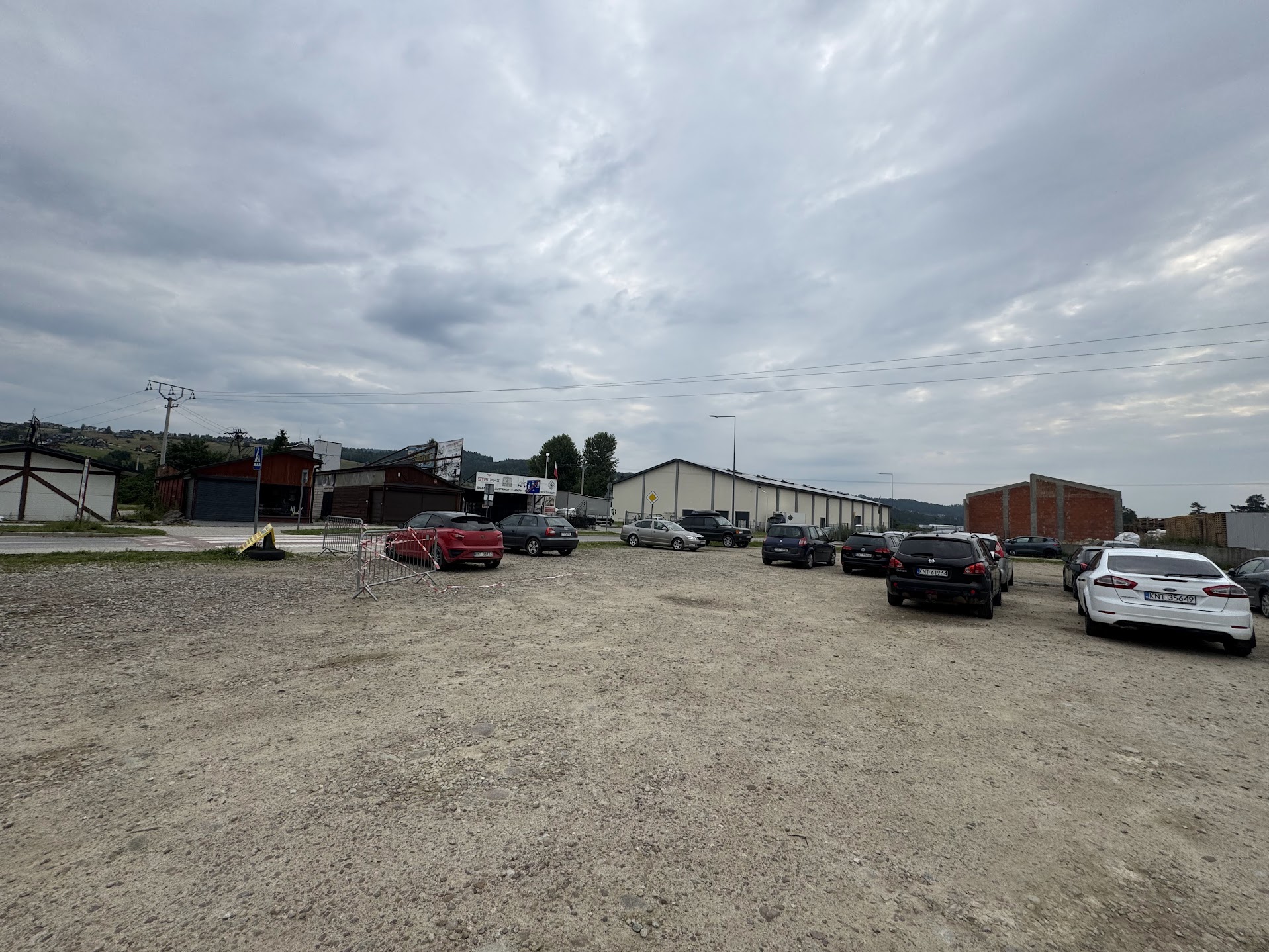 Fotografia przedstawia żwirowy parking przemysłowy z kilkoma zaparkowanymi samochodami, halami magazynowymi i ogrodzeniami