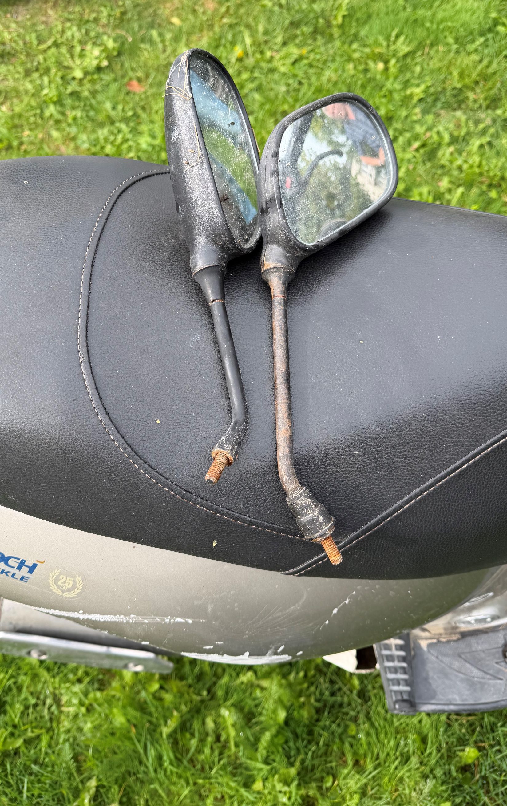 Widok czarnego siedzenia motoroweru z dwoma, odkconymi od kierownicy lusterkami wstecznymi.