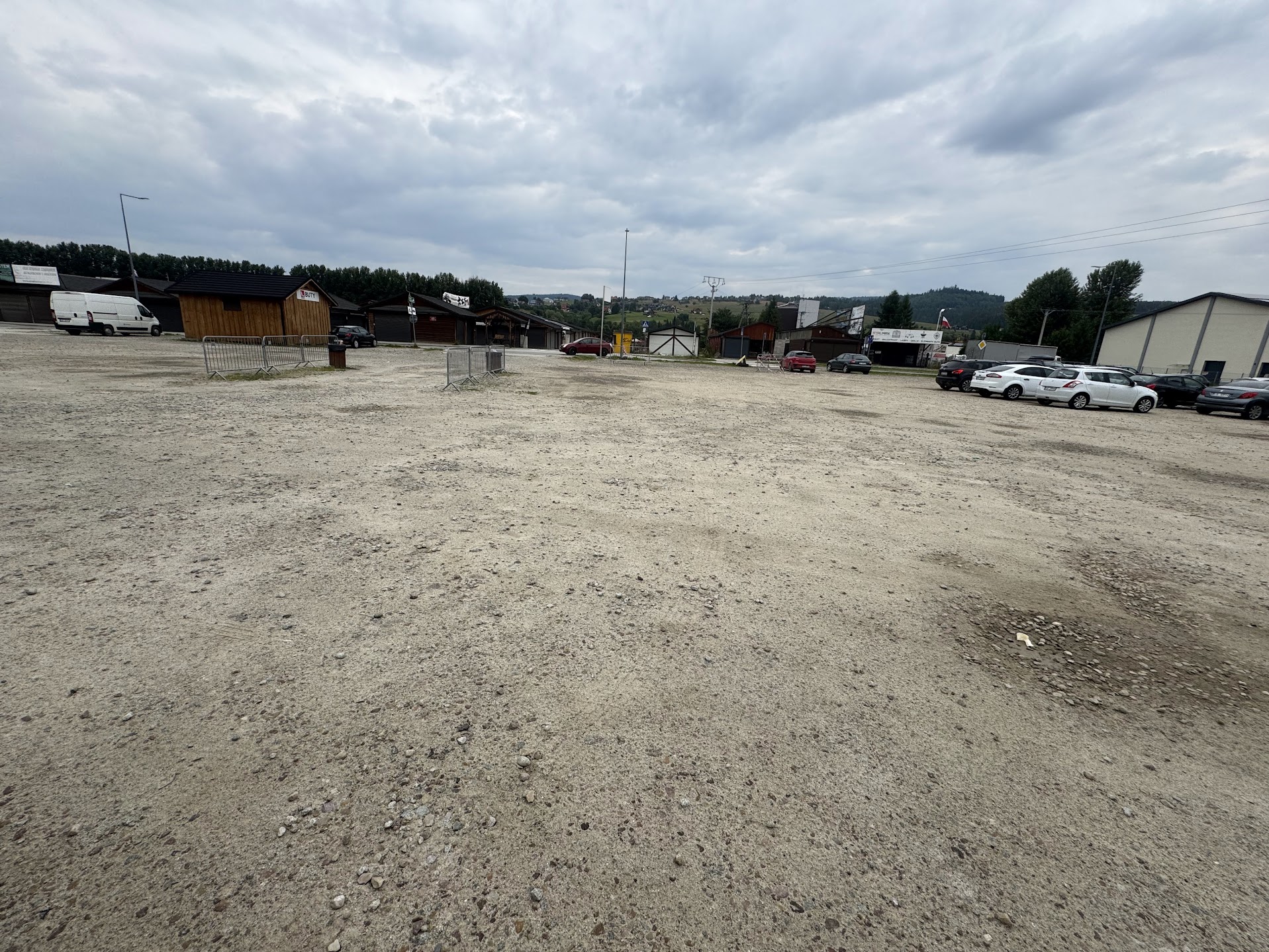 Fotografia przedstawia rozległy, żwirowy plac parkingowo-magazynowy. W tle widoczne są hale, drewniane zabudowania.