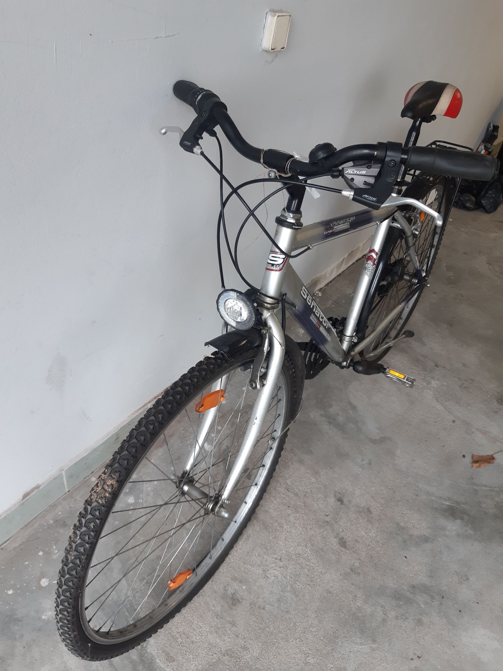 Srebrny rower Senator z czarną kierownicą, okrągłą przednią lampką, oponą z bieżnikiem i widoczną manetką Shimano Altus.