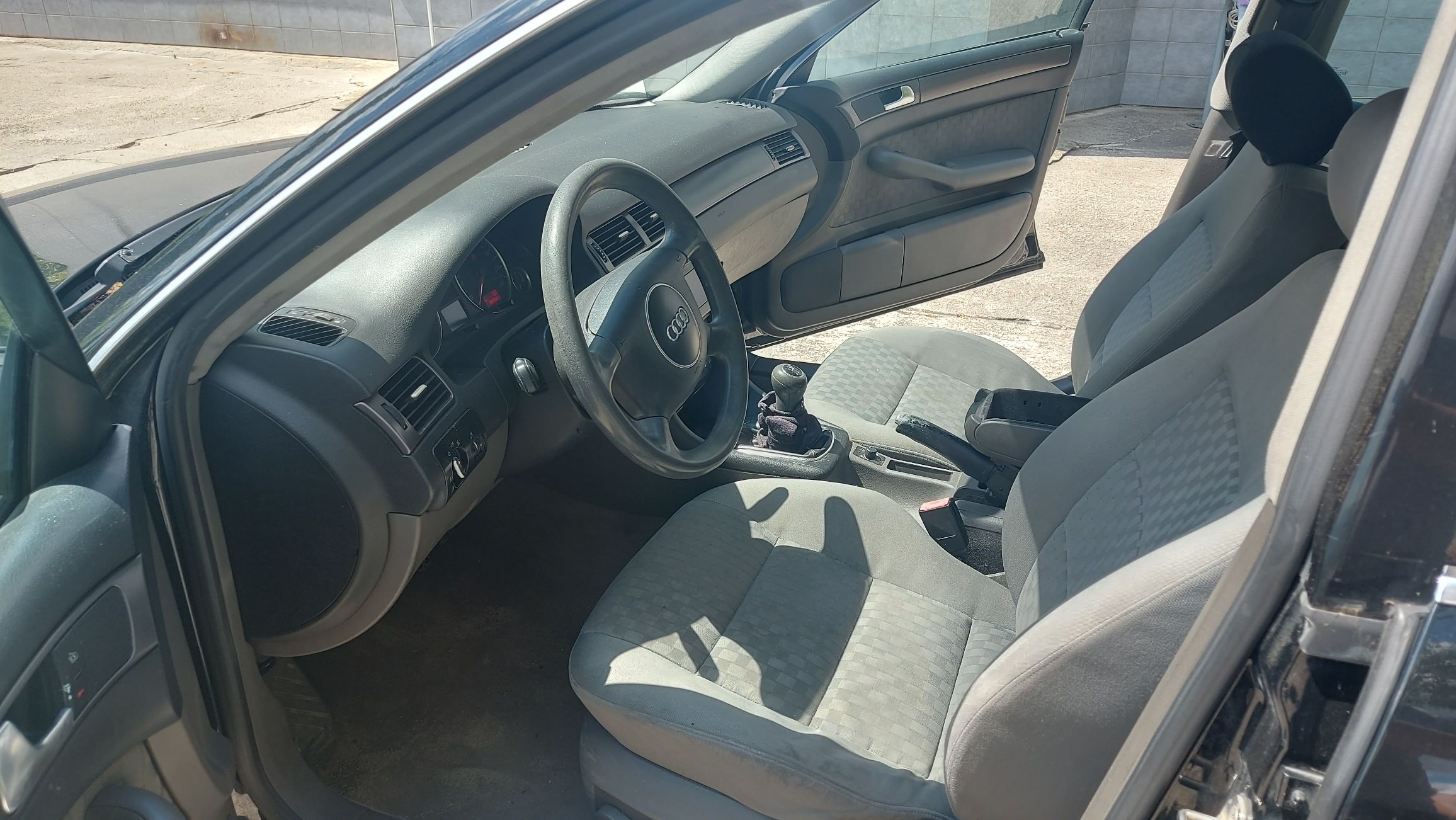 Wnętrze Audi A6 C5 od strony kierowcy. Widoczna kierownica, manualna skrzynia biegów oraz szara, materiałowa tapicerka foteli.