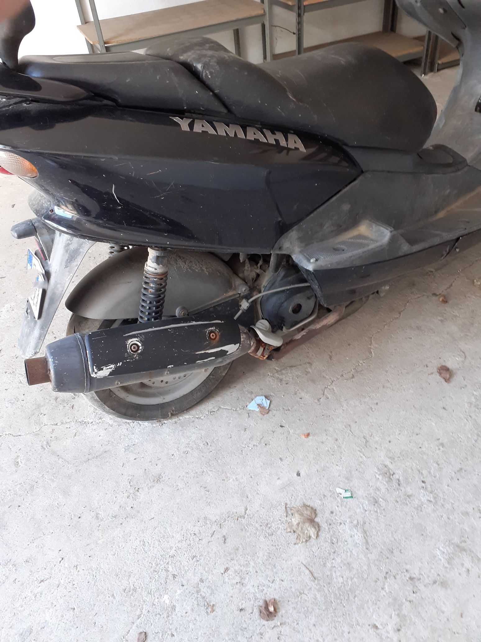 Tył czarnego motoroweru Yamaha Majesty, pokazujące porysowany tłumik wydechowy, tylne koło z amortyzatorem oraz fragment kanapy.