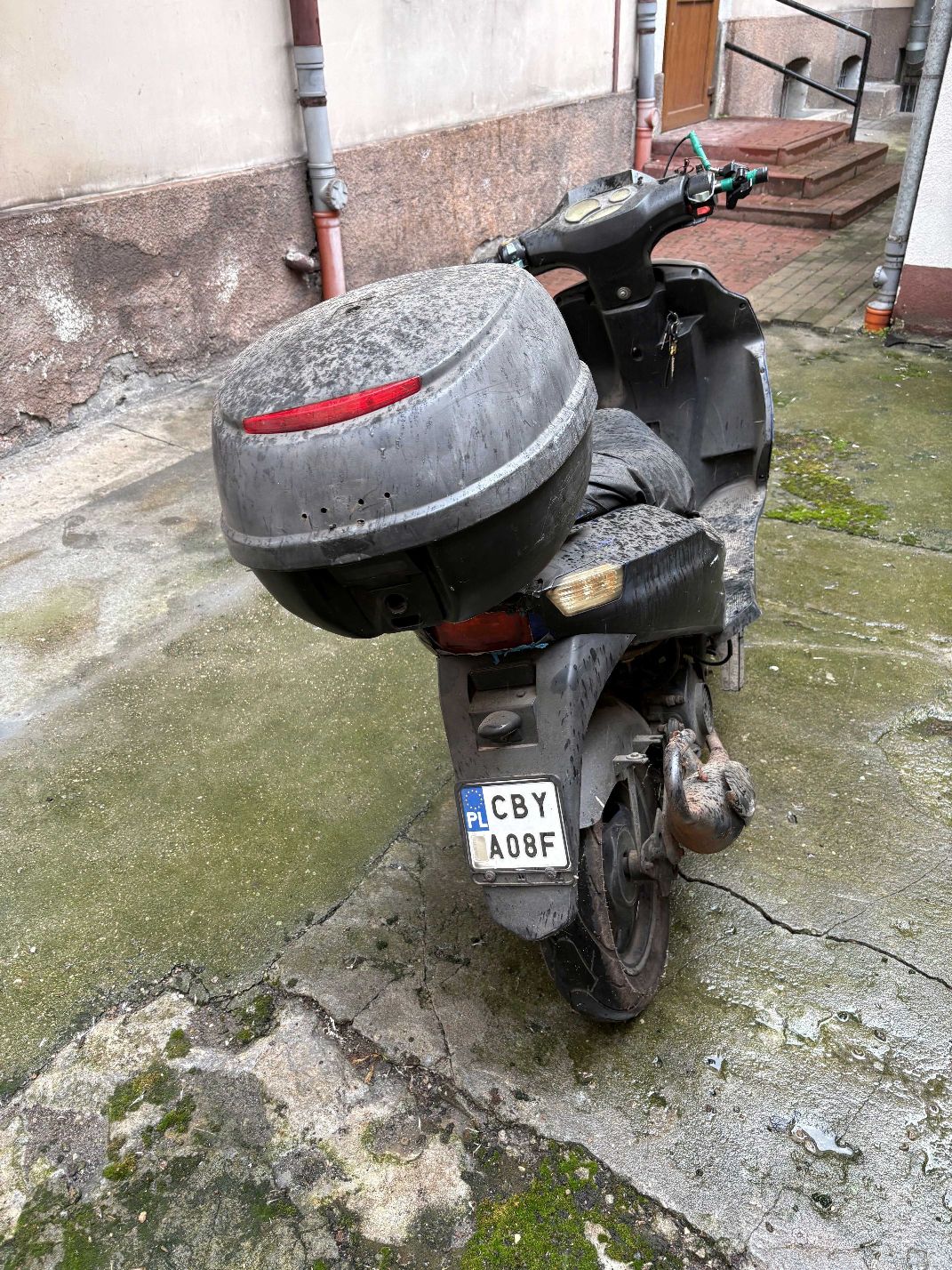 Motorower koloru czarnego wraz z szarym kufrem , widok tyłu pojazdu.