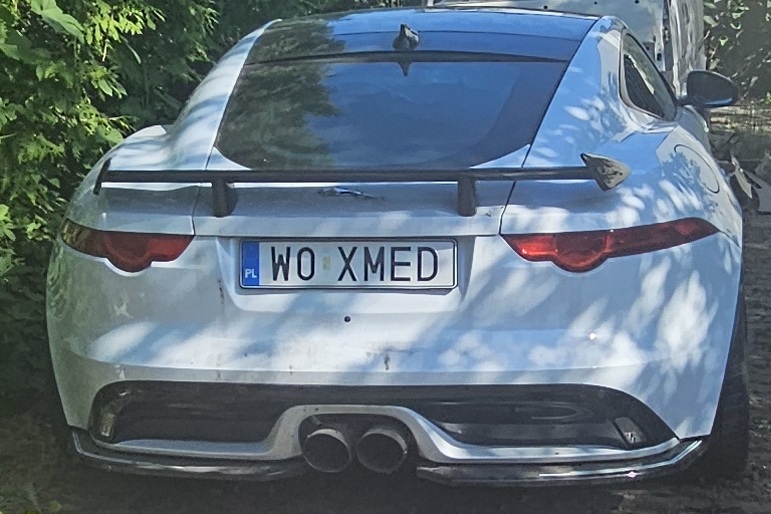 Tył białego jaguara, smukłe lampy, sportowy zderzak oraz centralnie umieszczony czarny spoiler. Dynamiczny charakter samochodu.
