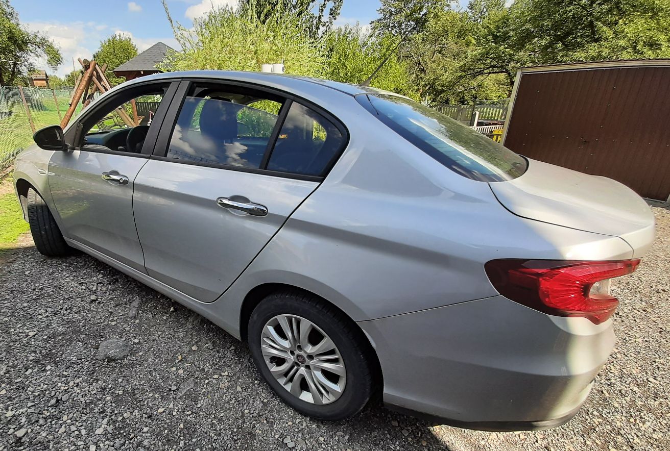 Srebrny FIAT TIPO ujęty od lewej strony. Samochód ma bagażnik, widoczne tylne światło, boczne lusterko, klamki drzwi.