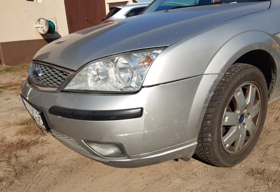 Srebrny Ford Mondeo ze zarysowanym przednim zderzakiem, widoczny od strony reflektora i koła.